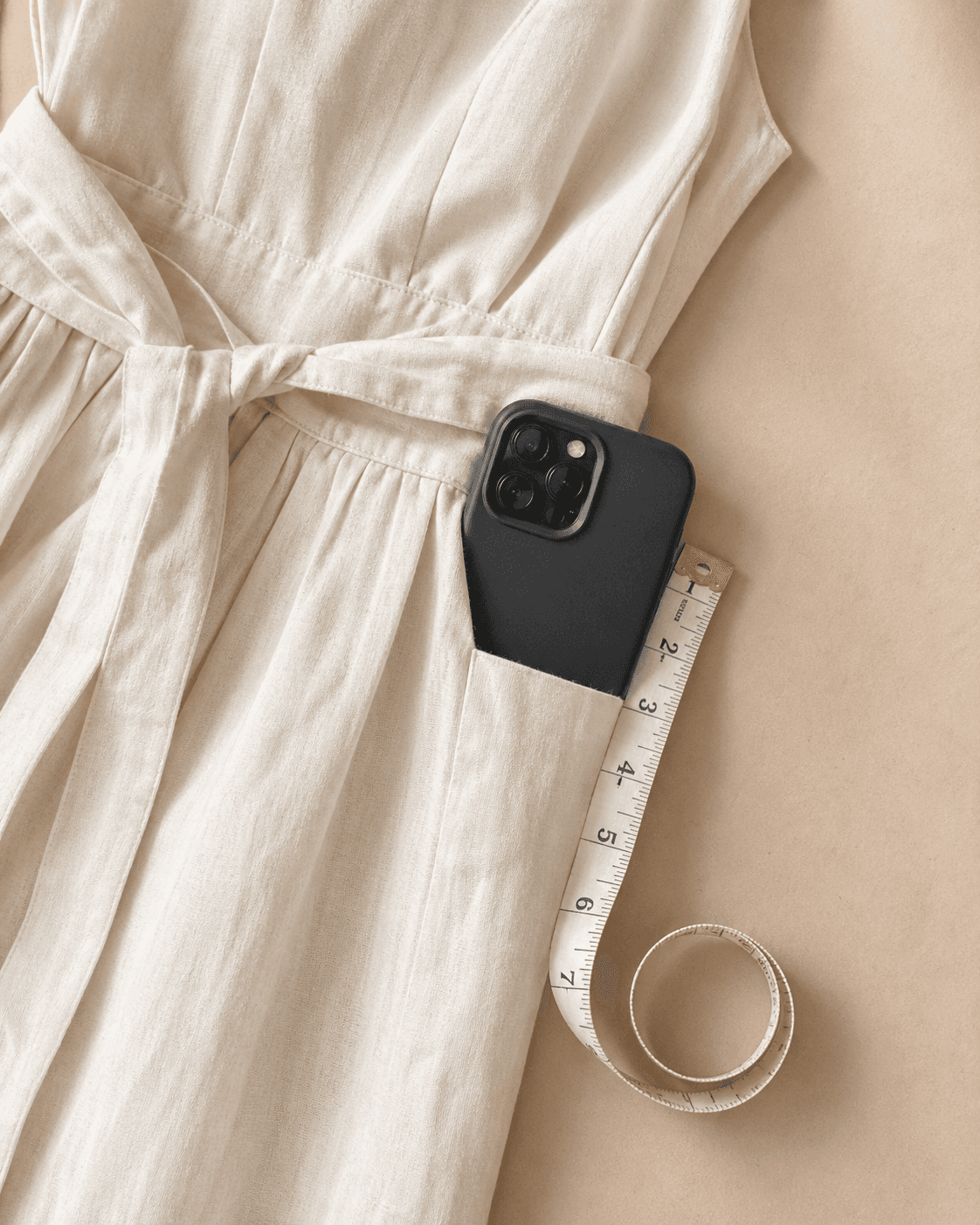 A cream linen dress with a smartphone tucked into a deep side-seam pocket, alongside a vintage cloth measuring tape showing the seven-inch depth required for a phone-fit pocket.
